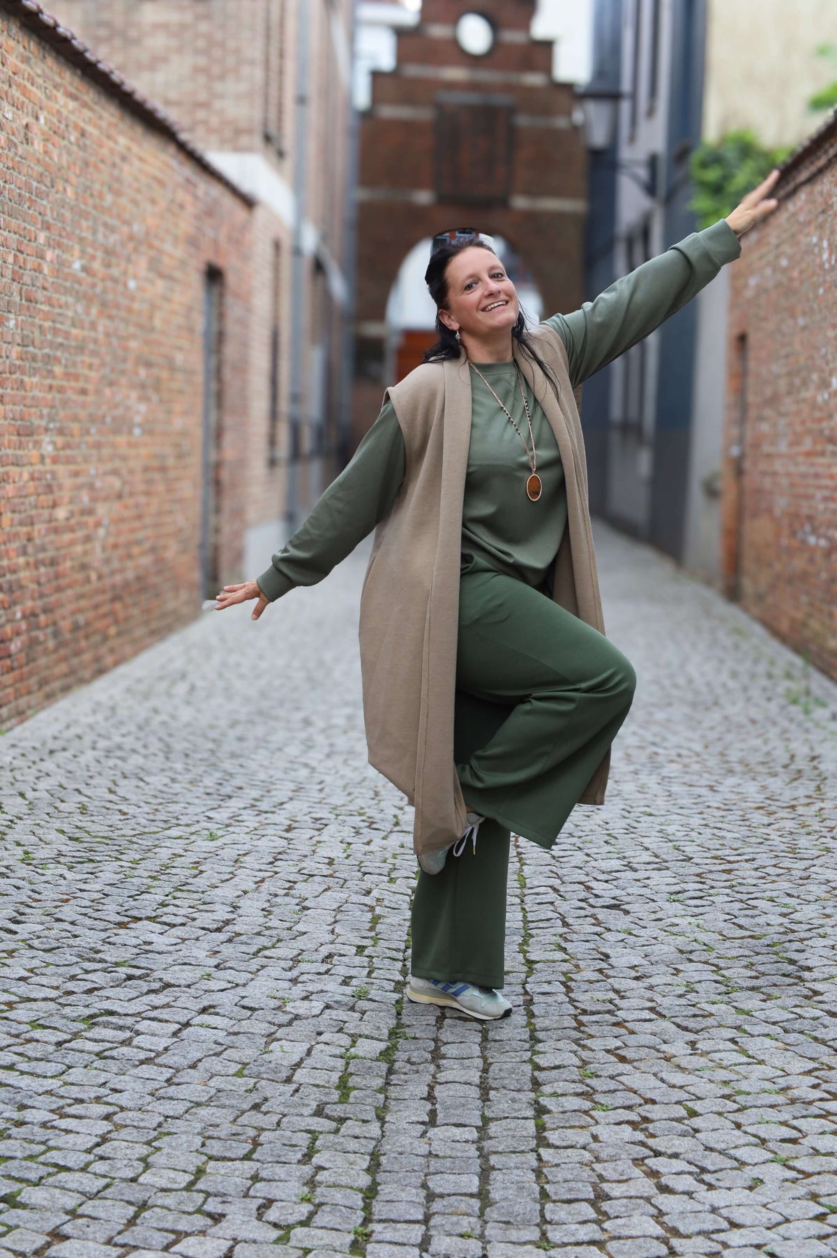 Vrouw in een lange beige gilet met een groene outfit poseert in een smalle straat met een architecturale achtergrond