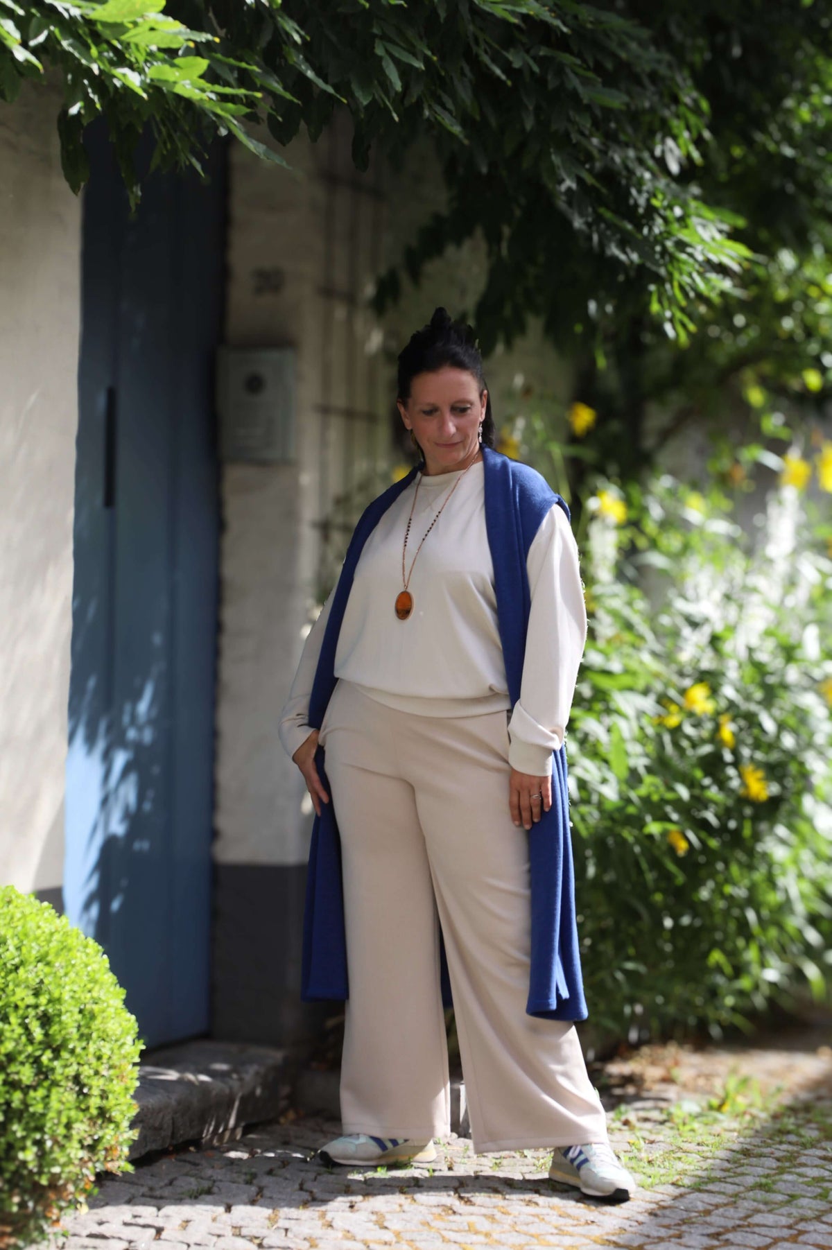 Vrouw in een blauwe gilet poseert zelfverzekerd buiten naast een muur, gekleed in een casual beige outfit.