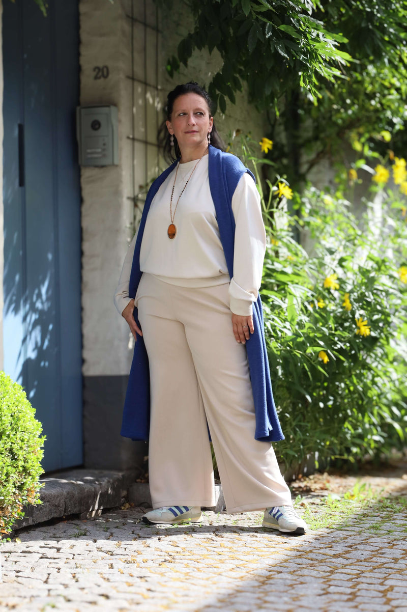 Vrouw in een blauwe gilet poseert zelfverzekerd buiten naast een muur, gekleed in een casual beige outfit.