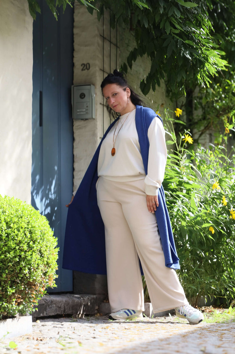 Vrouw in een blauwe gilet poseert zelfverzekerd buiten naast een muur, gekleed in een casual beige outfit.