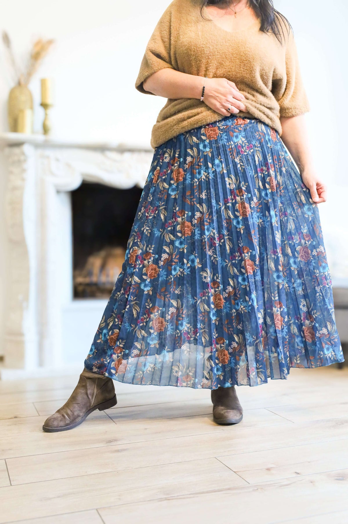 Vrouw draagt een plooienrok met bloemenprint in blauw, gecombineerd met een bruin truitje en enkellaarsjes