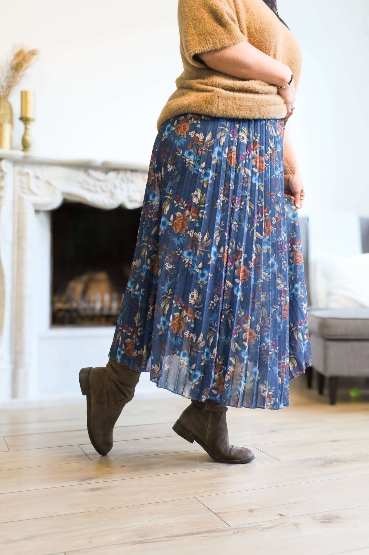 Vrouw draagt een plooienrok met bloemenprint in blauw, gecombineerd met een bruin truitje en enkellaarsjes