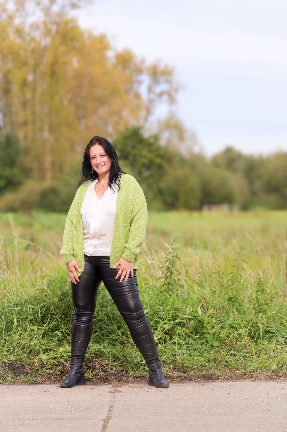 Vrouw draagt een gebreide cardigan in lime van Terra di Siena, gecombineerd met een witte blouse en leren broek, in een buitenomgeving.