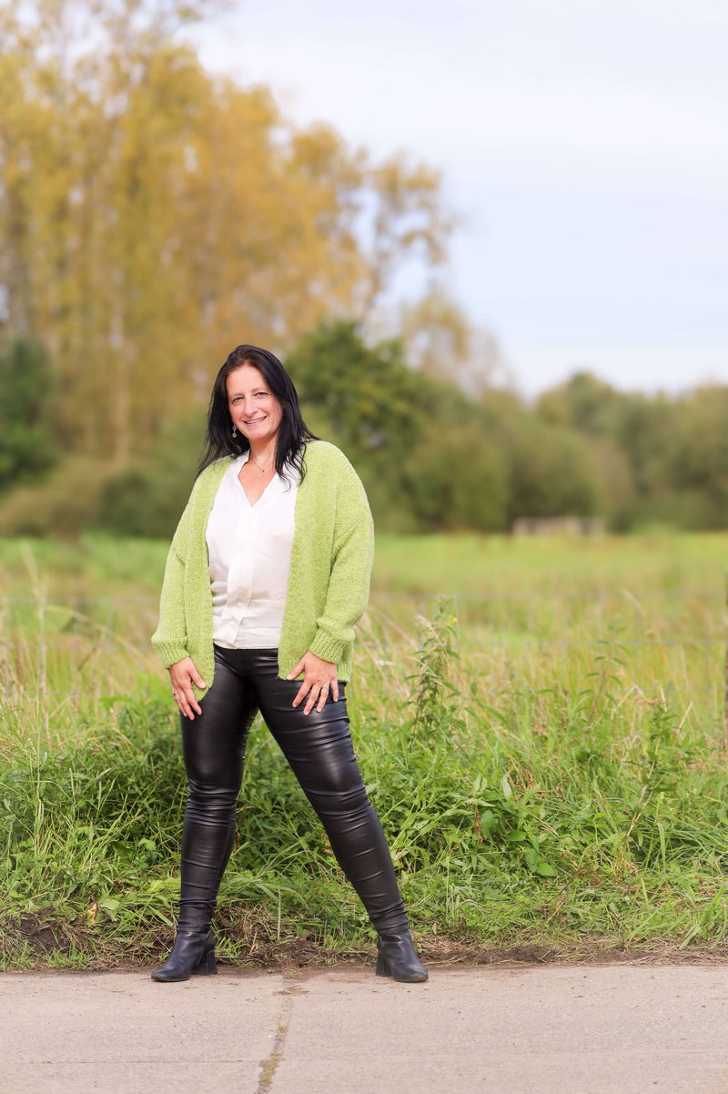 Vrouw draagt een gebreide cardigan in lime van Terra di Siena, gecombineerd met een witte blouse en leren broek, in een buitenomgeving.