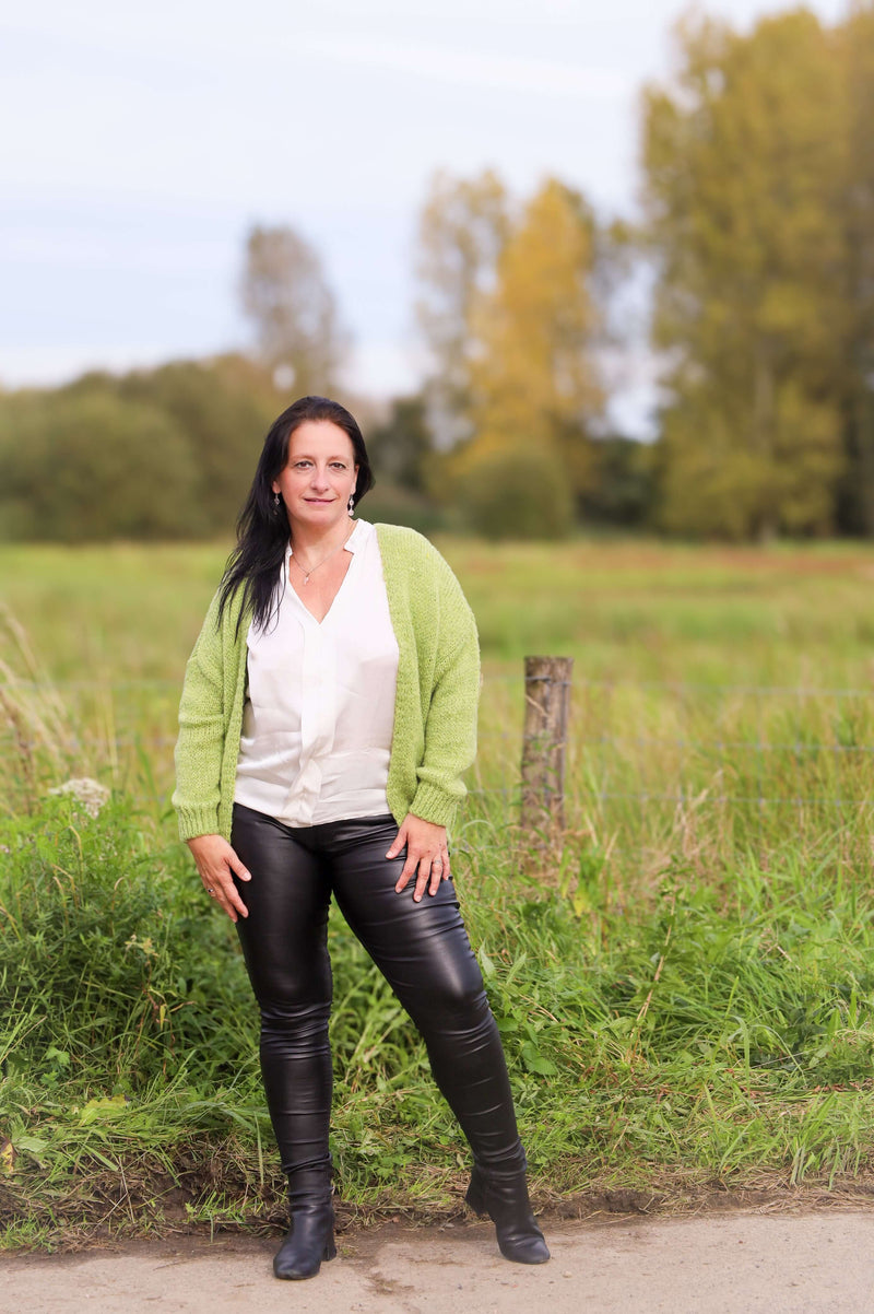 Vrouw draagt een gebreide cardigan in lime van Terra di Siena, gecombineerd met een witte blouse en leren broek, in een buitenomgeving.