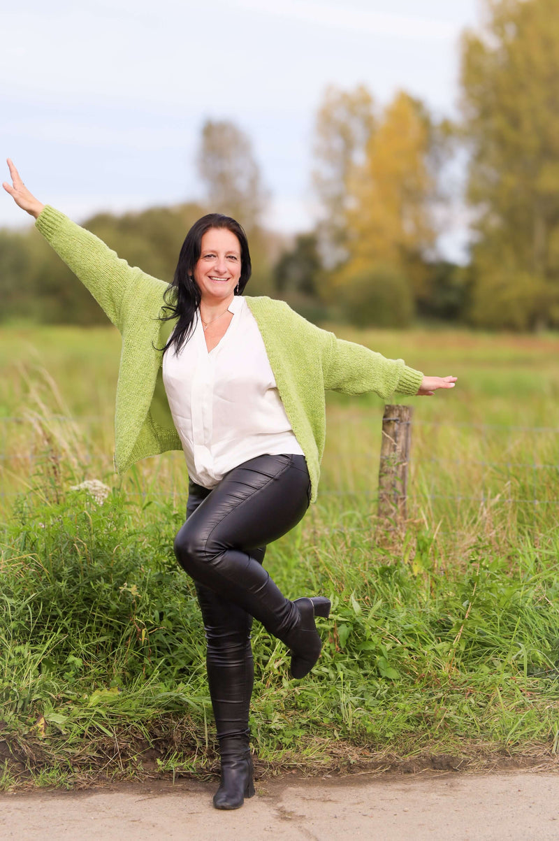 Vrouw draagt een gebreide cardigan in lime van Terra di Siena, gecombineerd met een witte blouse en leren broek, in een buitenomgeving.