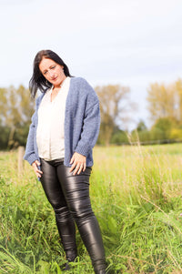 Vrouw draagt een gebreide cardigan in jeansblauw van Terra di Siena, gecombineerd met een witte blouse en leren broek, in een buitenomgeving.