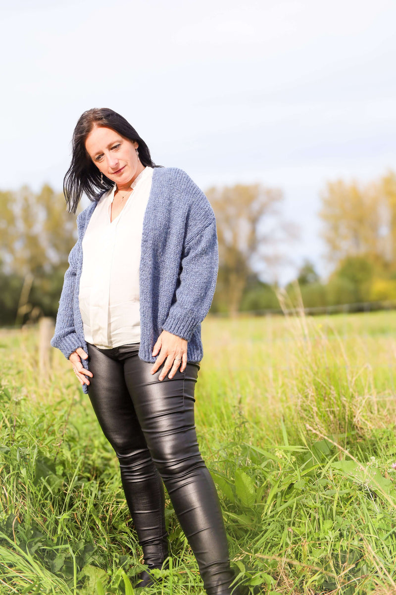 Vrouw draagt een gebreide cardigan in jeansblauw van Terra di Siena, gecombineerd met een witte blouse en leren broek, in een buitenomgeving.