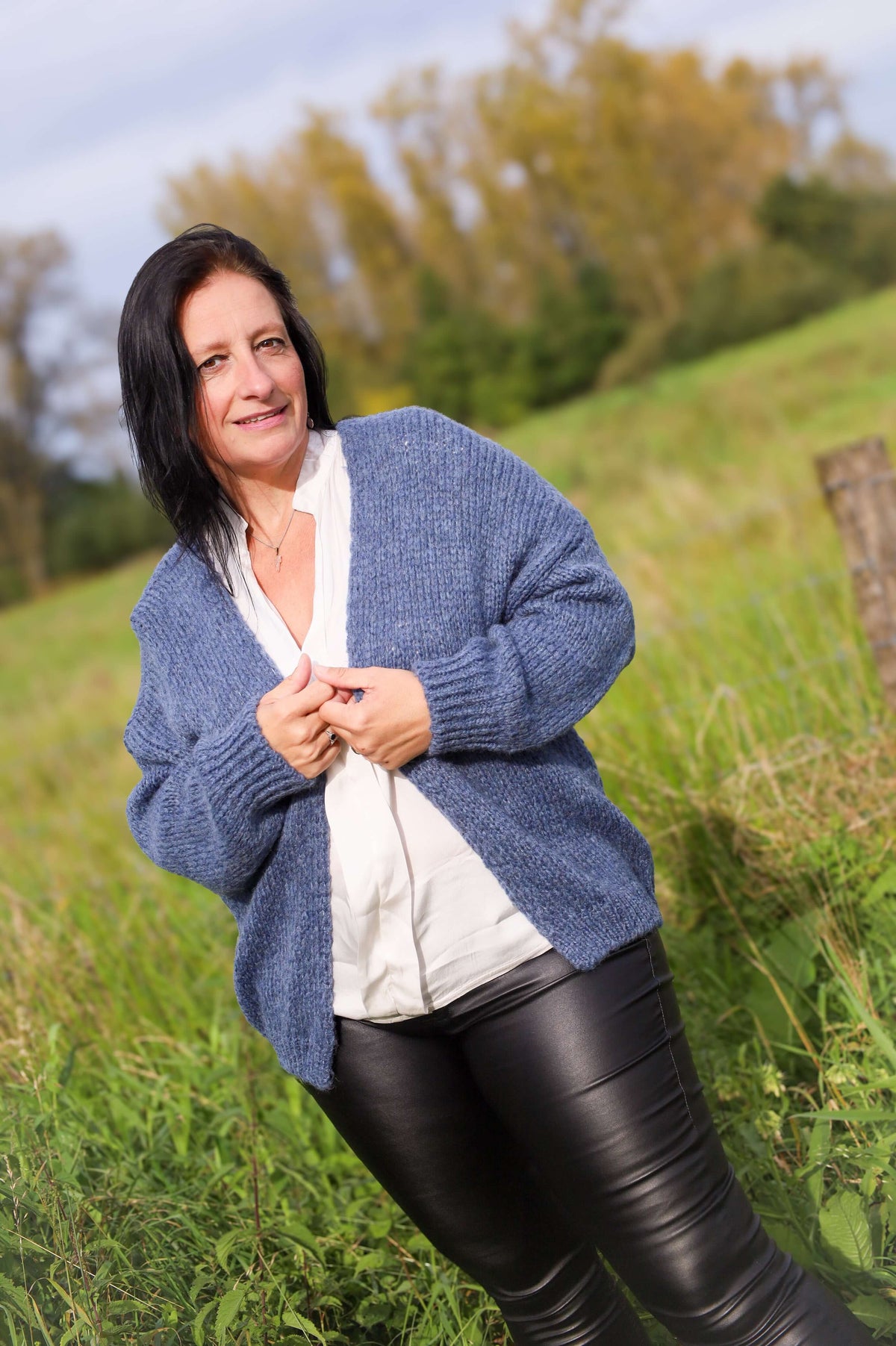 Vrouw draagt een gebreide cardigan in jeansblauw van Terra di Siena, gecombineerd met een witte blouse en leren broek, in een buitenomgeving.