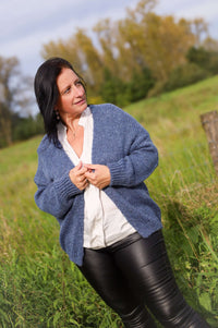 Vrouw draagt een gebreide cardigan in jeansblauw van Terra di Siena, gecombineerd met een witte blouse en leren broek, in een buitenomgeving.