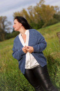 Vrouw draagt een gebreide cardigan in jeansblauw van Terra di Siena, gecombineerd met een witte blouse en leren broek, in een buitenomgeving.