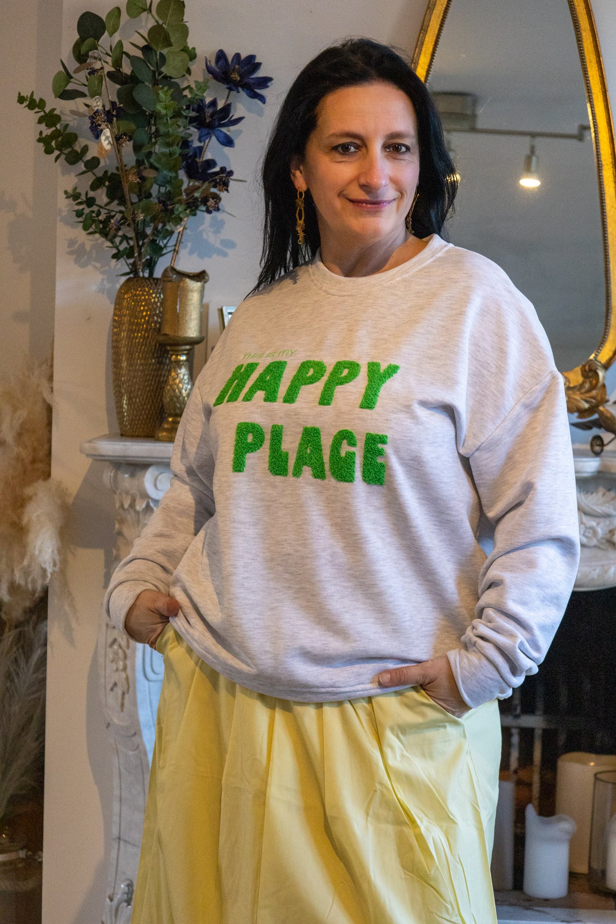 Model in grijze sweater met groene tekst 'HAPPY PLACE' en gele plissérok.
