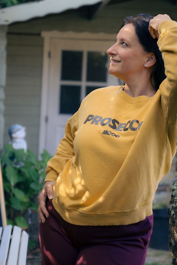 Vrouw draagt mosterdgele Dolci Giorni sweater met ‘Prosecco Mood’-print – comfortabele damesmode bij She&Me Duffel.