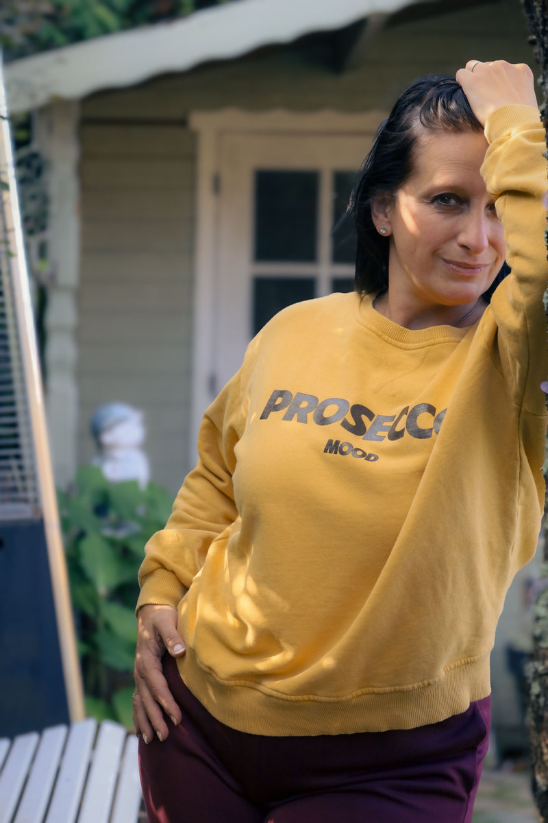 Vrouw draagt mosterdgele Dolci Giorni sweater met ‘Prosecco Mood’-print – comfortabele damesmode bij She&Me Duffel.