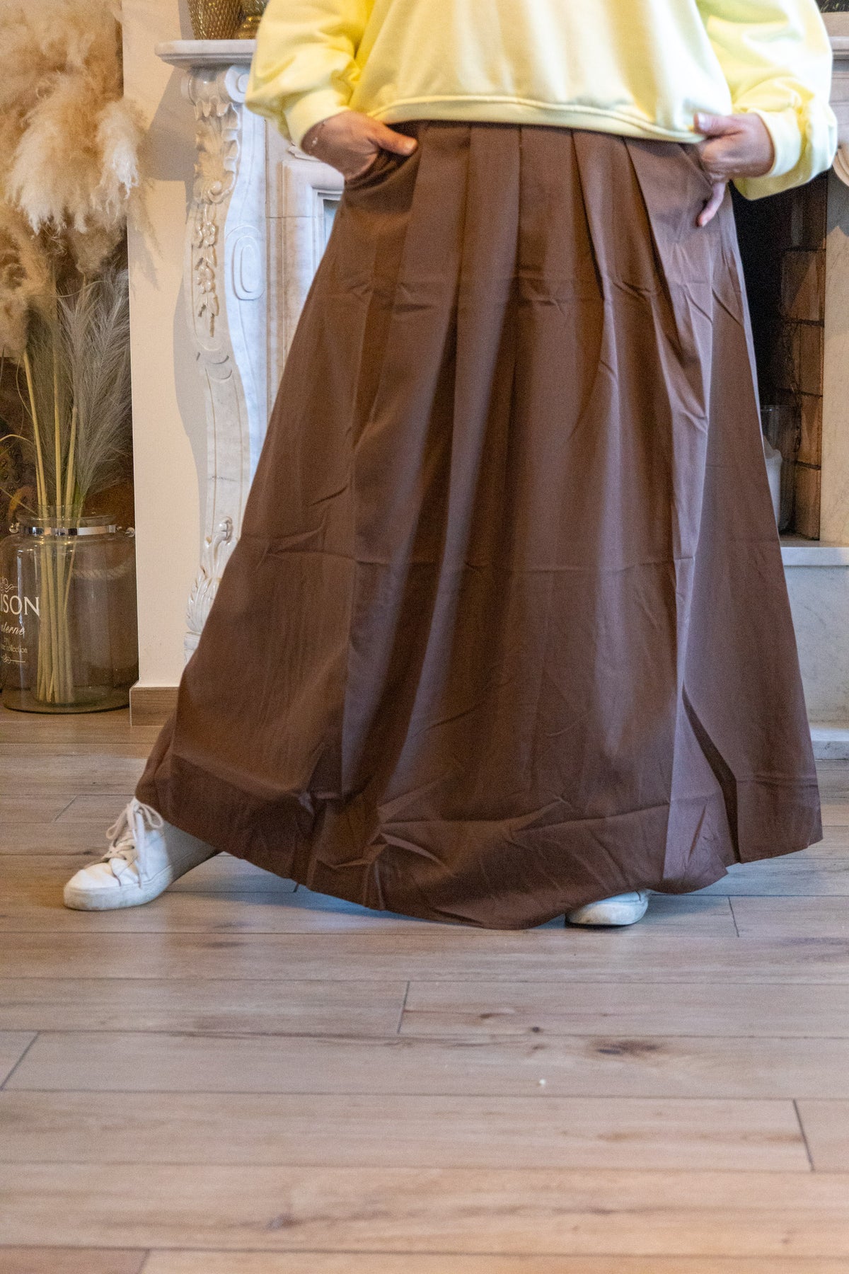 Elegante lange bruine rok met plooien, stijlvol gecombineerd met een gele trui en witte sneakers.