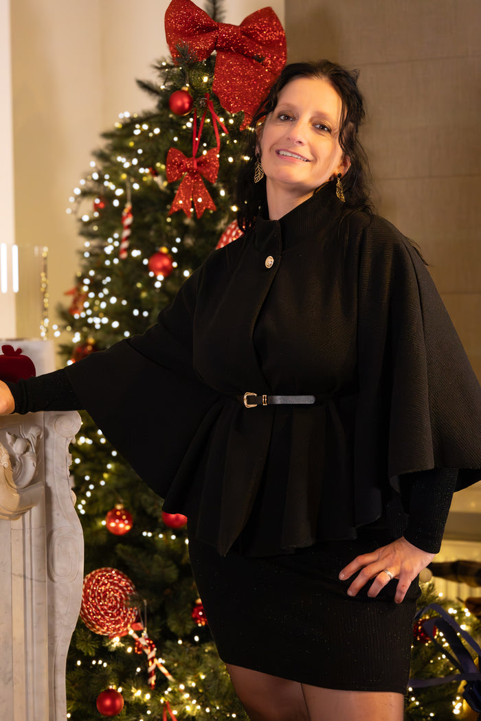 Zwart cape-achtig vest met strikceintuur gedragen door vrouw, feestelijke uitstraling met kerstdecoratie.