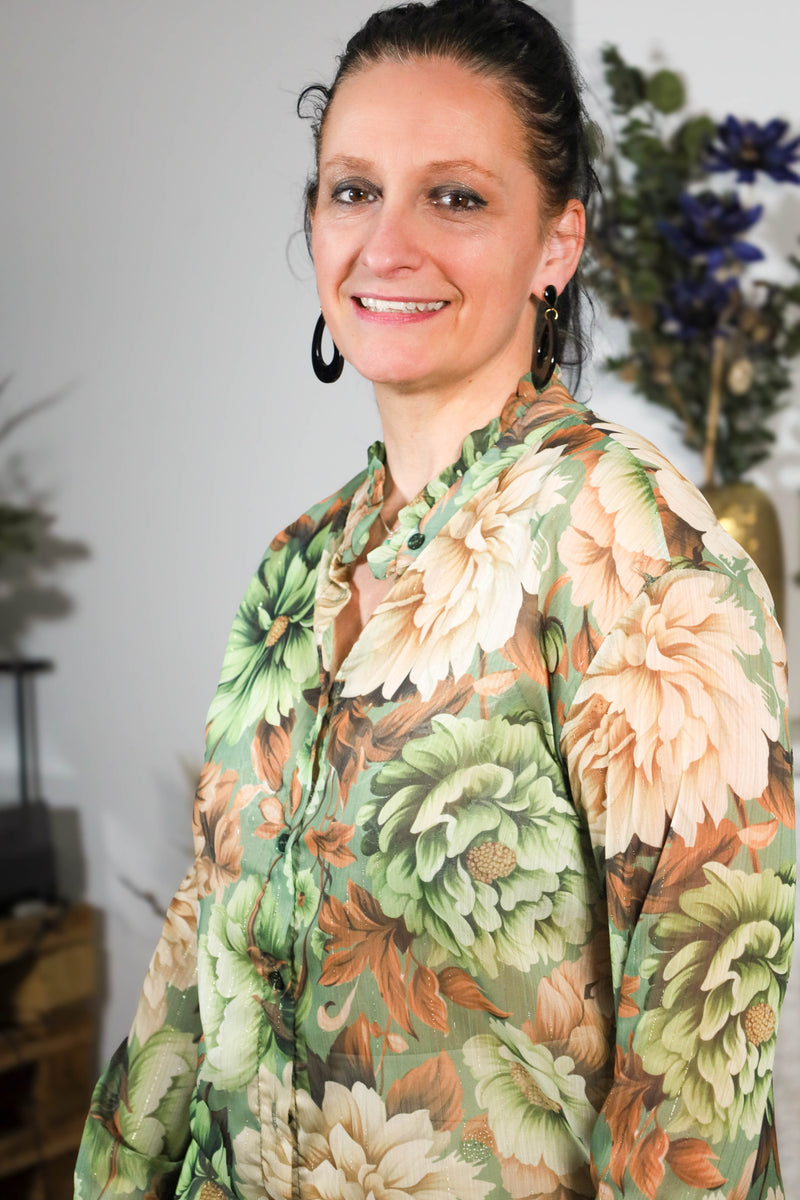 Vrouw draagt een transparante blouse met bloemenprint in groene en beige tinten, gecombineerd met een subtiele kraag en natuurlijke uitstraling