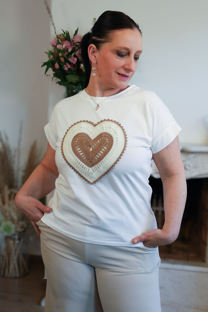 Vrouw draagt een wit T-shirt met een geborduurd hart in beige tinten, gecombineerd met een lichte broek