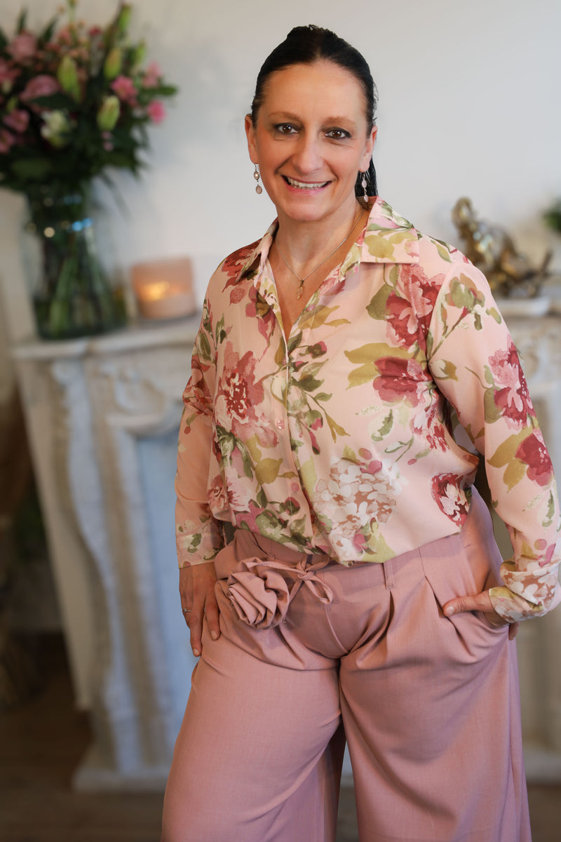 Vrouw draagt een transparante blouse met romantische bloemenprint, gecombineerd met een roze pantalon.