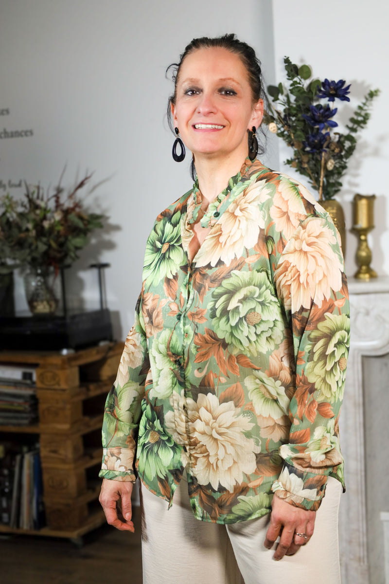 Vrouw draagt een transparante blouse met bloemenprint in groene en beige tinten, gecombineerd met een subtiele kraag en natuurlijke uitstraling